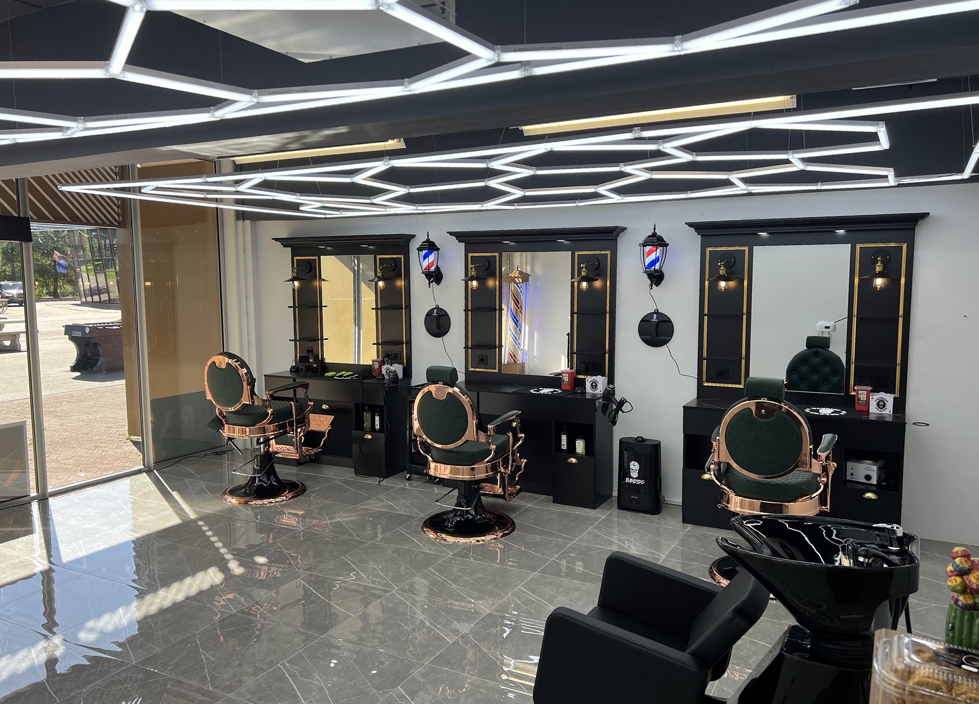 De salon van binnen: drie kappersstoelen met koperen frames, gouden spiegels en een plafond van zeshoekige lichtlijnen