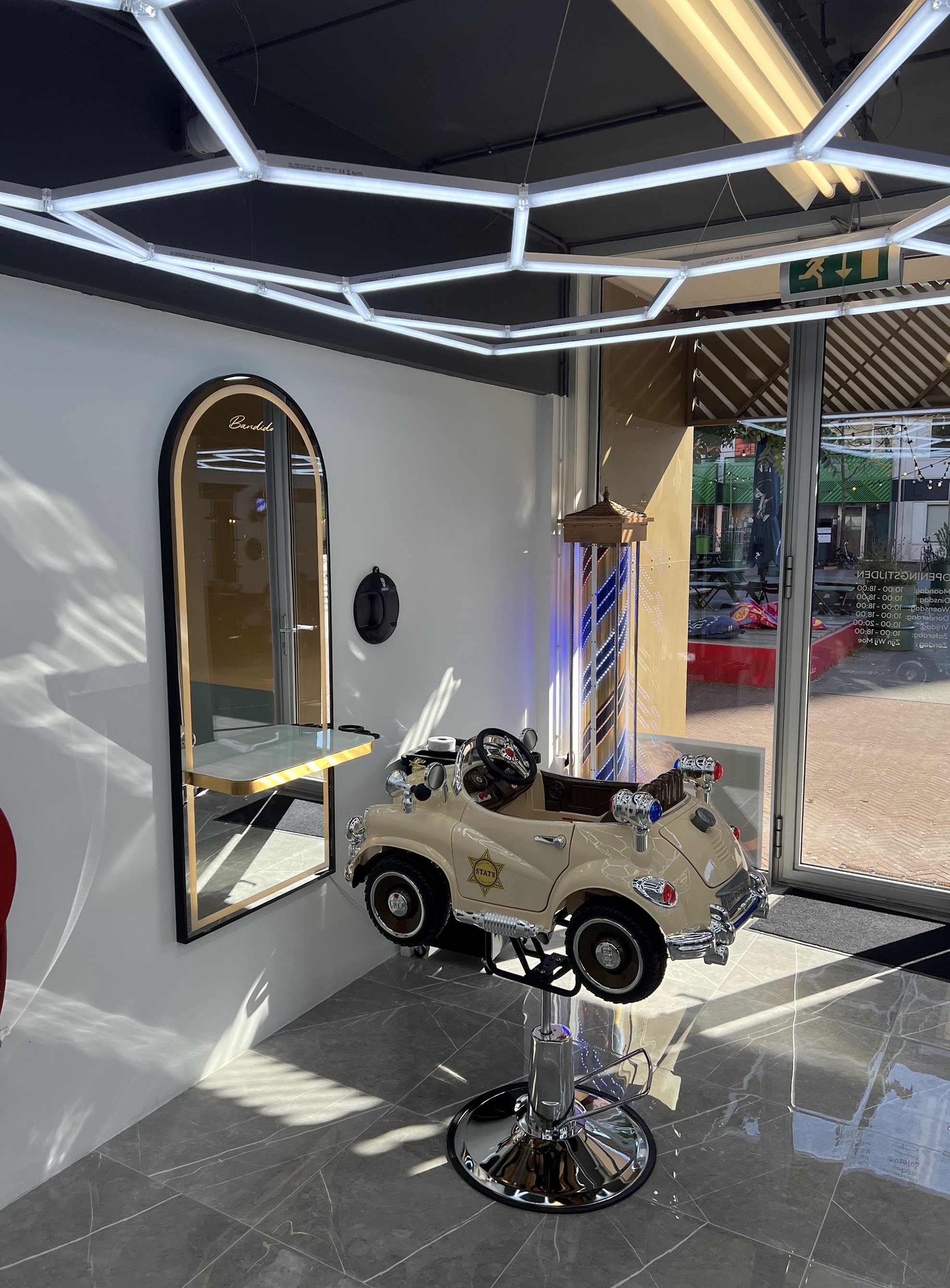 De kinderstoel in de vorm van een crèmekleurige auto, naast een gouden boogspiegel en een barberpaal